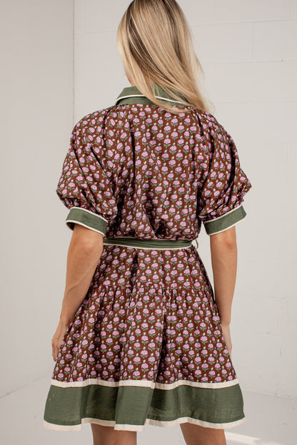 A woman viewed from behind wearing a short puff-sleeve dress with an allover floral print in brown, pink, and green, featuring a matching green waist tie and a green hem with cream piping.