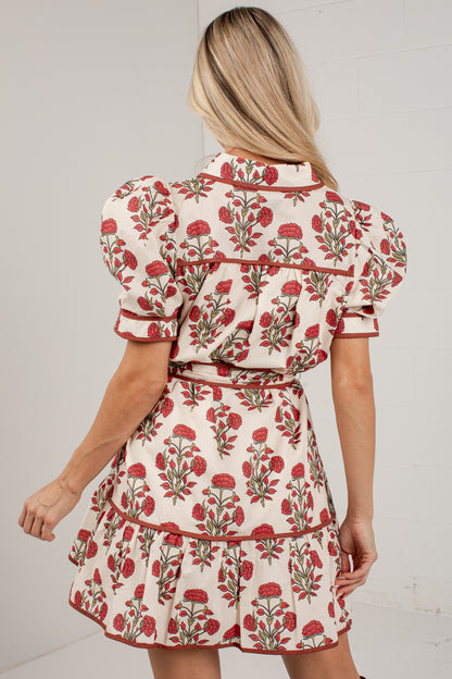 A woman shown from the back wearing a cream-colored mini dress with a red and green floral pattern, puffed short sleeves, and a ruffled waist.