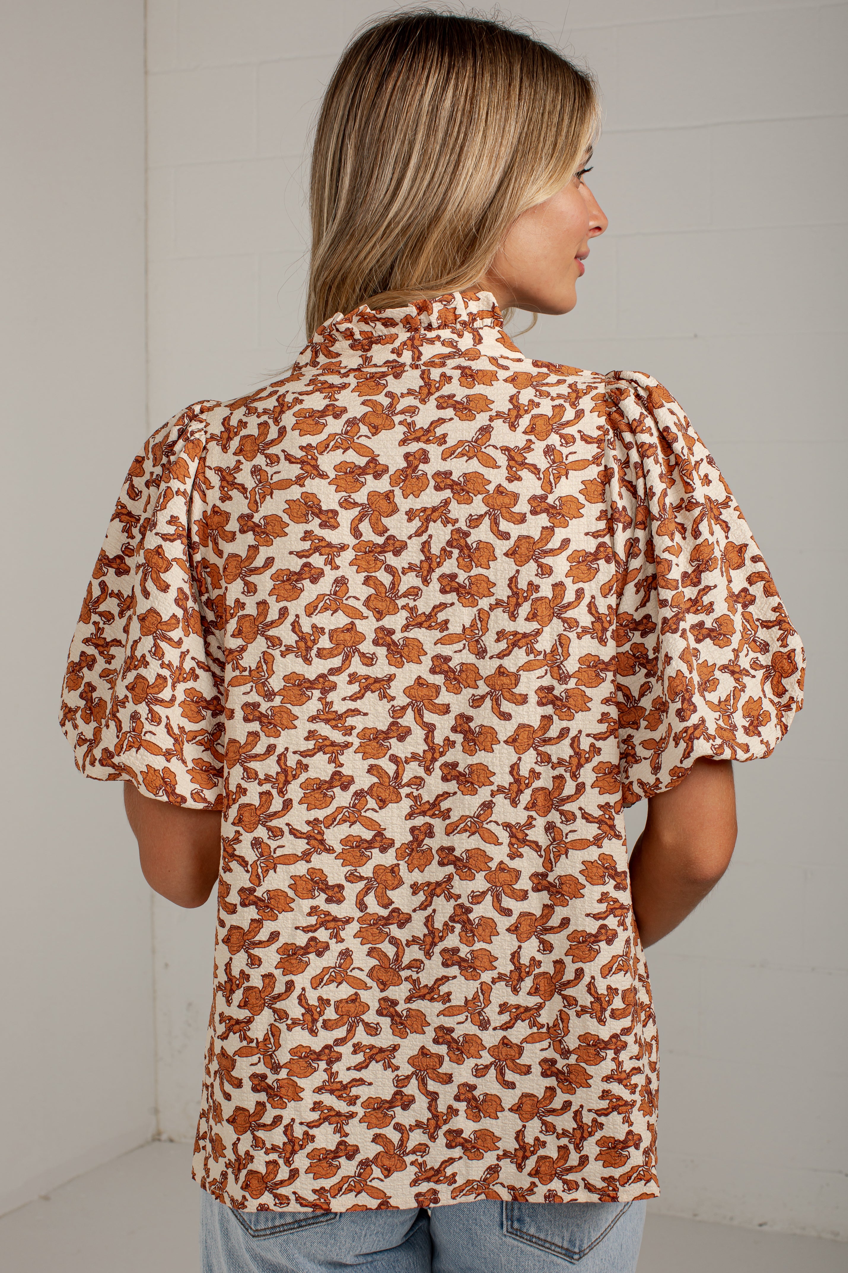 A woman shown from the back wearing a short-sleeve blouse with puff sleeves and an allover brown floral print on a cream background, paired with light blue jeans against a white wall.