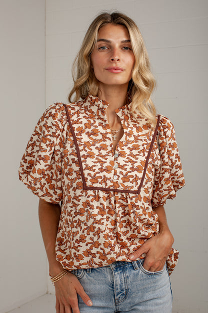 A woman wearing a short-sleeve blouse with a brown and beige abstract pattern and dark brown trim, paired with light blue jeans, standing against a light gray wall.