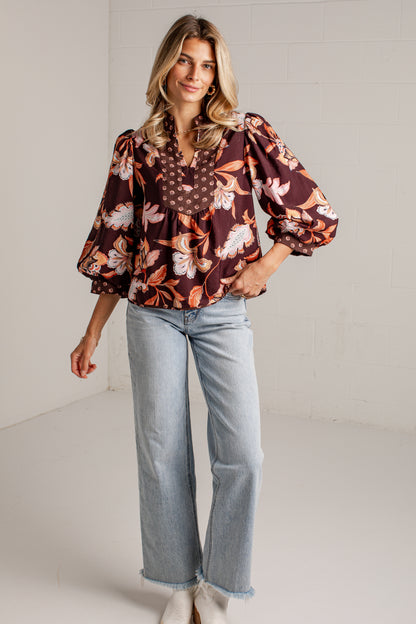 A woman modeling a brown floral blouse with puff sleeves and light-wash wide-leg jeans against a white studio wall.