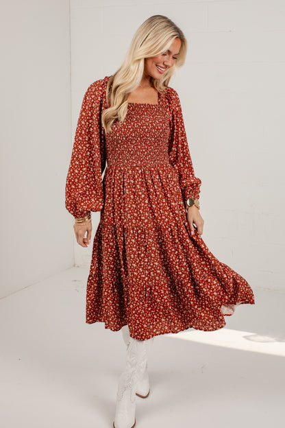 A woman modeling a rust-red midi dress with a white floral pattern, featuring long puff sleeves and a smocked bodice, paired with white lace-up boots in a bright studio setting.