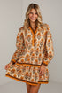 A woman modeling a long-sleeve tunic dress with a beige base, orange floral pattern, and brown trim against a light gray wall.