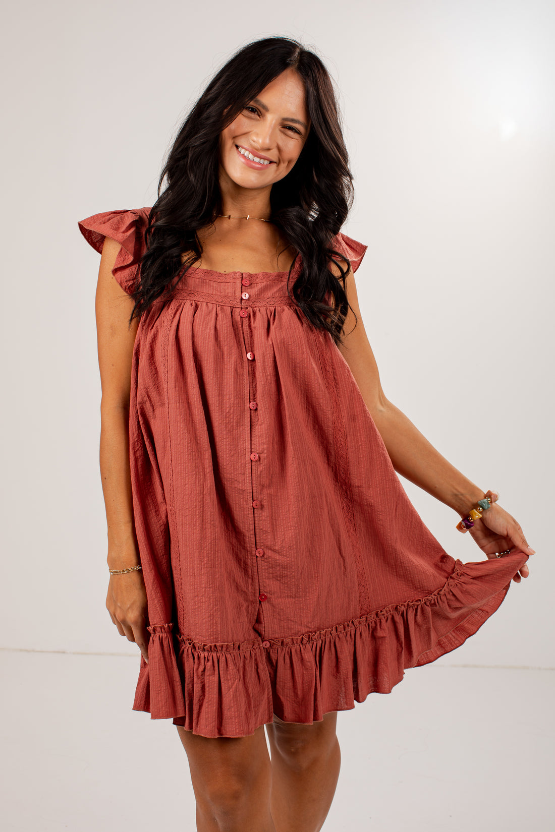 A woman modeling a rust-colored linen dress with ruffled sleeves and a button-down front, set against a plain white studio background.