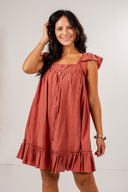 A woman modeling a terracotta-colored linen dress with ruffled shoulder straps, a button-down front, and a ruffled hem against a plain light gray background.