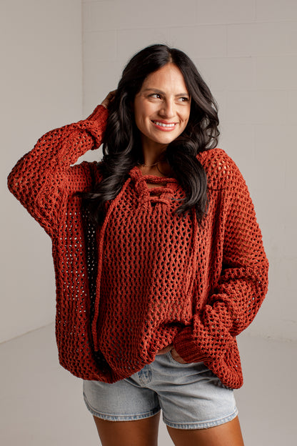 A woman wearing a rust-orange chunky knit crochet sweater with a tie-neck detail, paired with light-wash denim shorts, posing against a light gray brick wall.