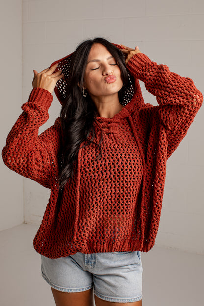 A woman wearing a chunky, open-knit rust-orange hoodie with drawstrings, posing with her hands on her hood in a bright studio setting.