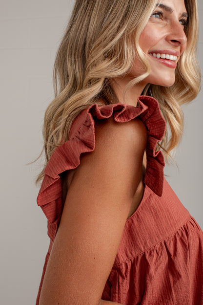 A close-up side profile of a smiling woman with blonde hair wearing a rust-colored sleeveless top with ruffled shoulder straps and a gathered waist, set against a plain light gray studio background.