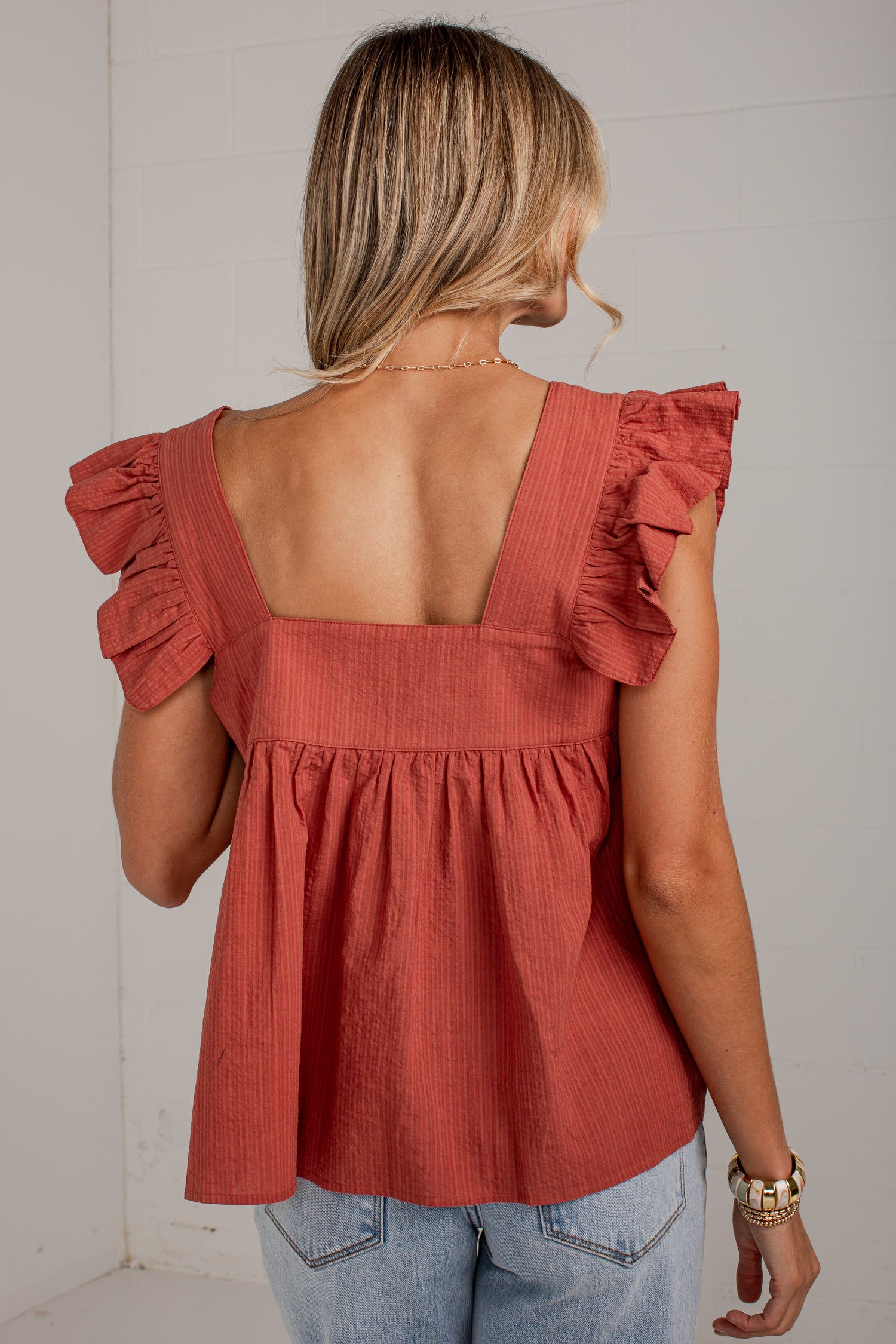 A woman shown from the back wearing a terracotta-colored ruffled top with a square open back and light-wash jeans, standing against a white wall.