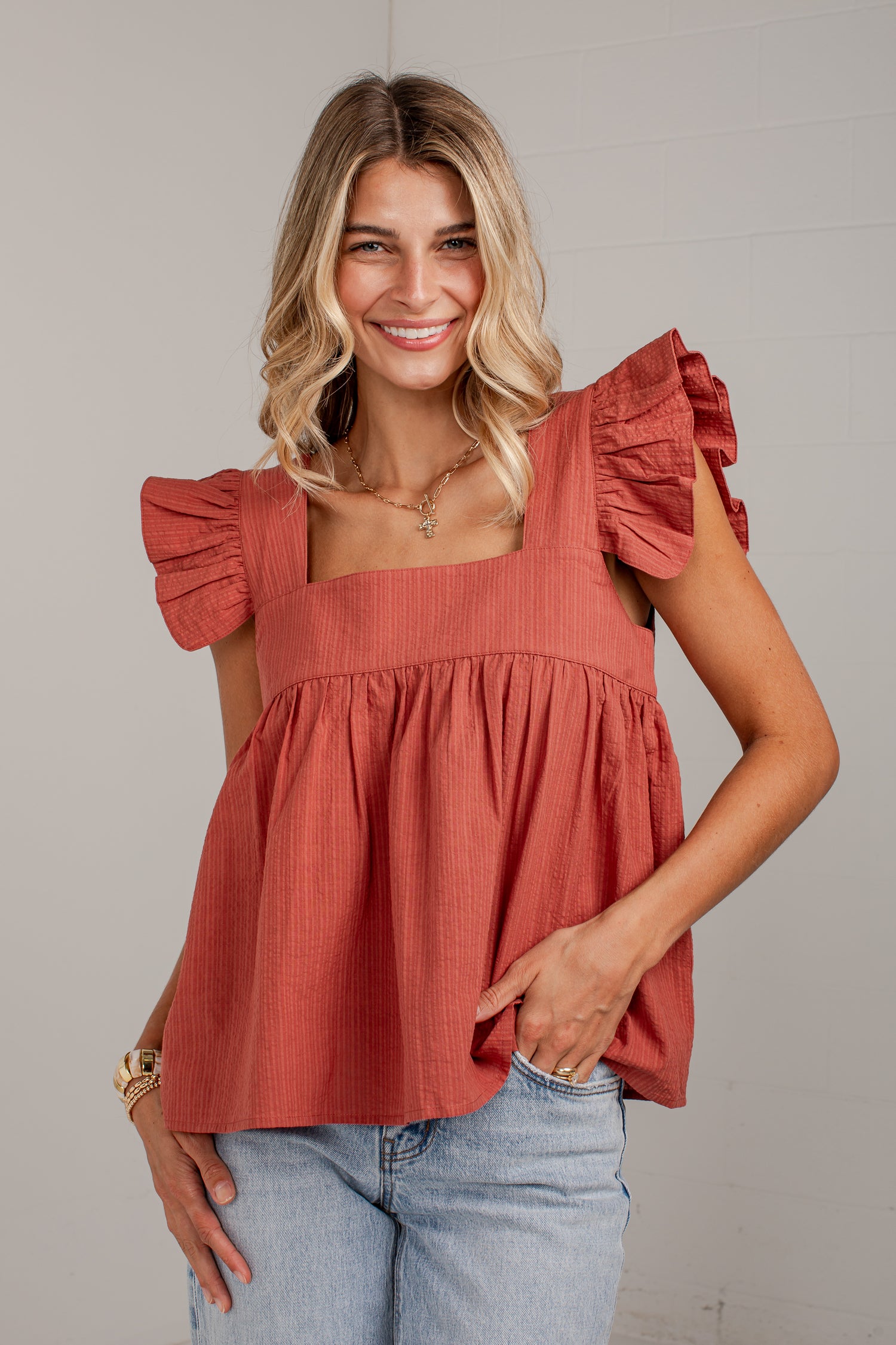 A woman wearing a terracotta-colored ruffled blouse with a square neckline and light-wash blue jeans, standing against a light gray wall.