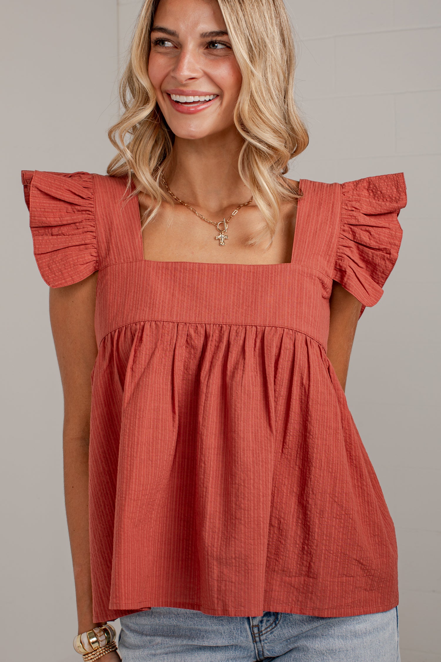 A woman wearing a terracotta-colored crinkle-textured top with ruffled cap sleeves and a square neckline, paired with light blue jeans.