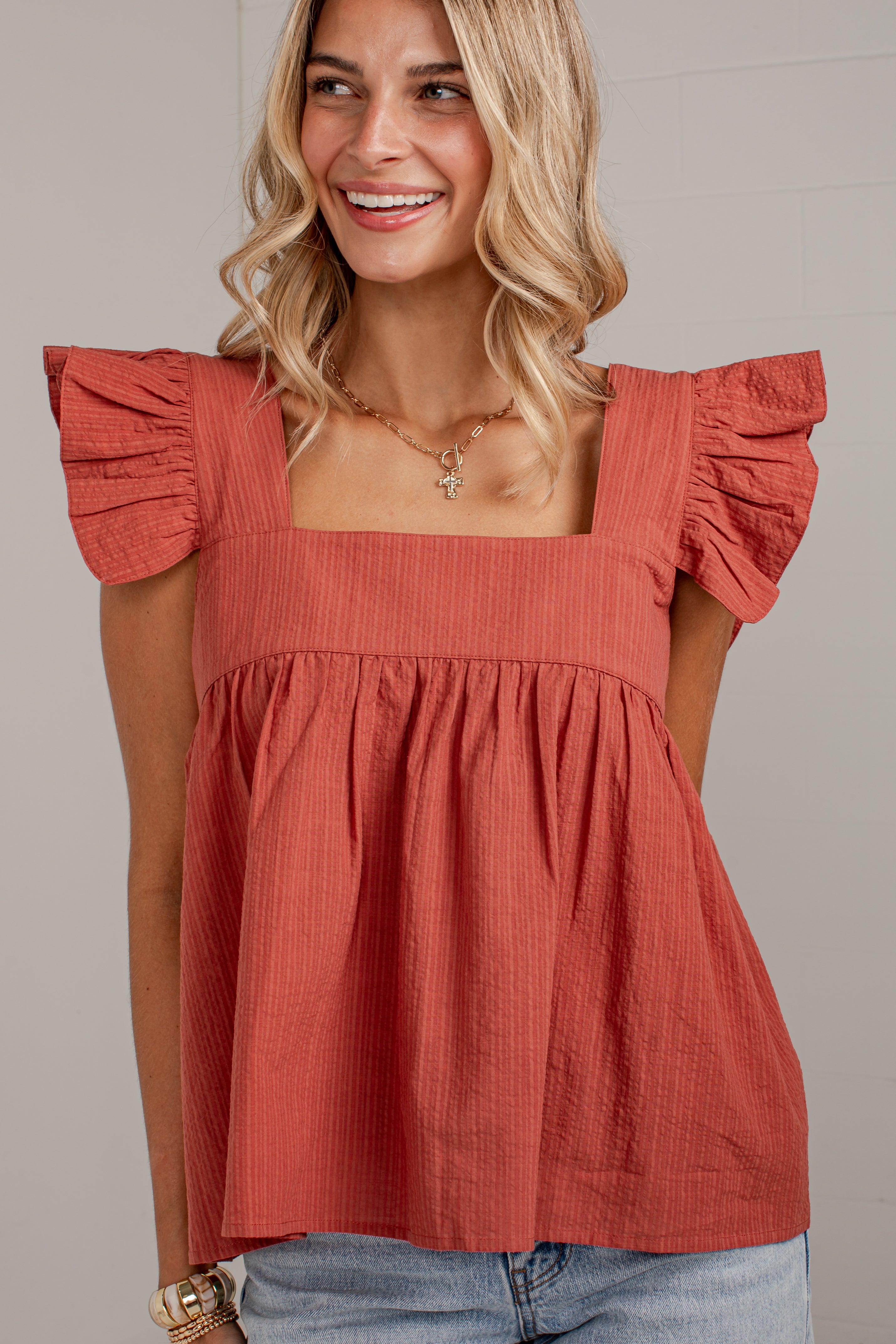 A woman wearing a terracotta-colored crinkle-textured top with ruffled cap sleeves and a square neckline, paired with light blue jeans.