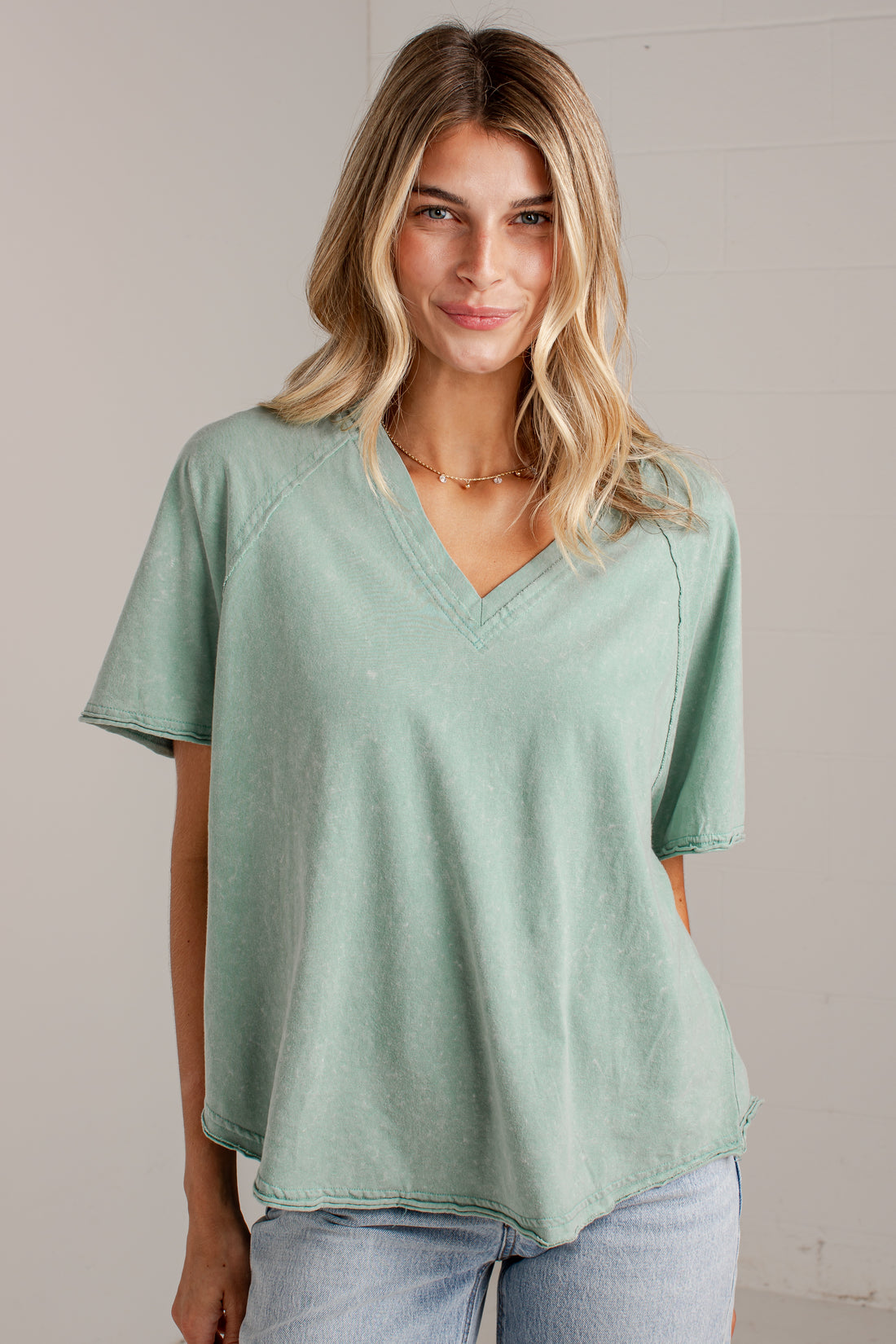 A woman wearing a light sage green V-neck short-sleeve top with a relaxed, slightly oversized fit and light wash blue jeans, standing against a neutral light gray wall.