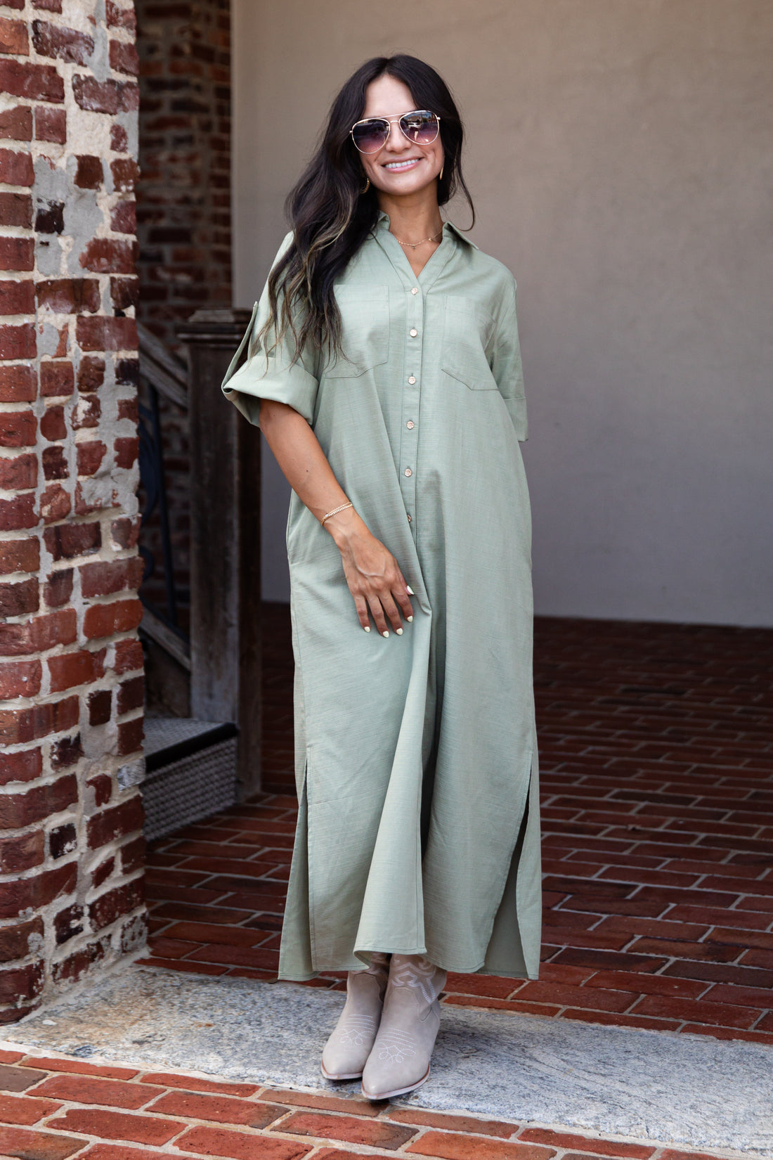 A woman wearing a sage green short-sleeve button-up maxi shirt dress, styled with sunglasses, a bracelet, and beige ankle boots, standing in a brick doorway.