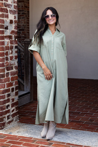 A woman wearing a sage green short-sleeve button-up maxi shirt dress, styled with sunglasses, a bracelet, and beige ankle boots, standing in a brick doorway.