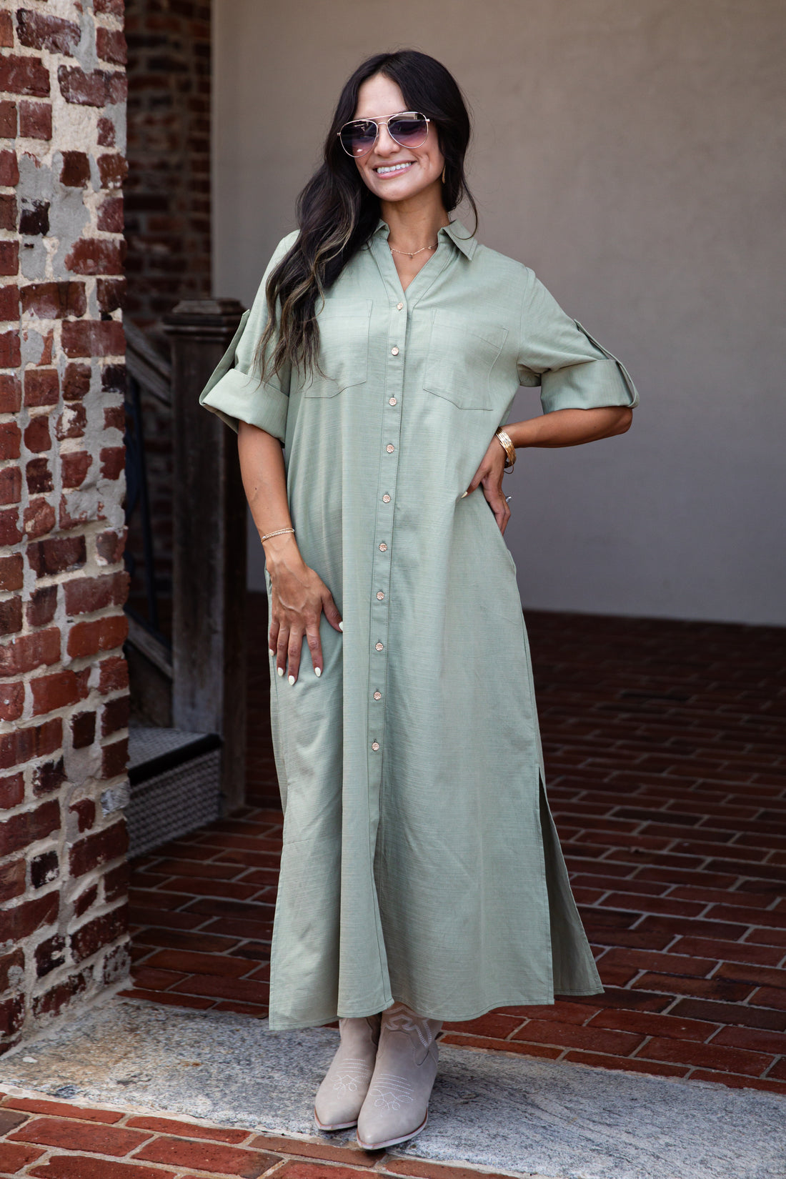 A woman wearing a sage green short-sleeve button-up maxi shirt dress, styled with beige ankle boots, sunglasses, and a gold watch, standing in front of a brick and stucco wall.