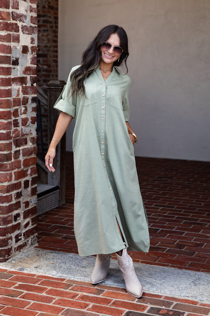 A woman wearing a sage green short-sleeve button-up maxi dress and light beige ankle boots, standing on a brick patio next to a brick column.