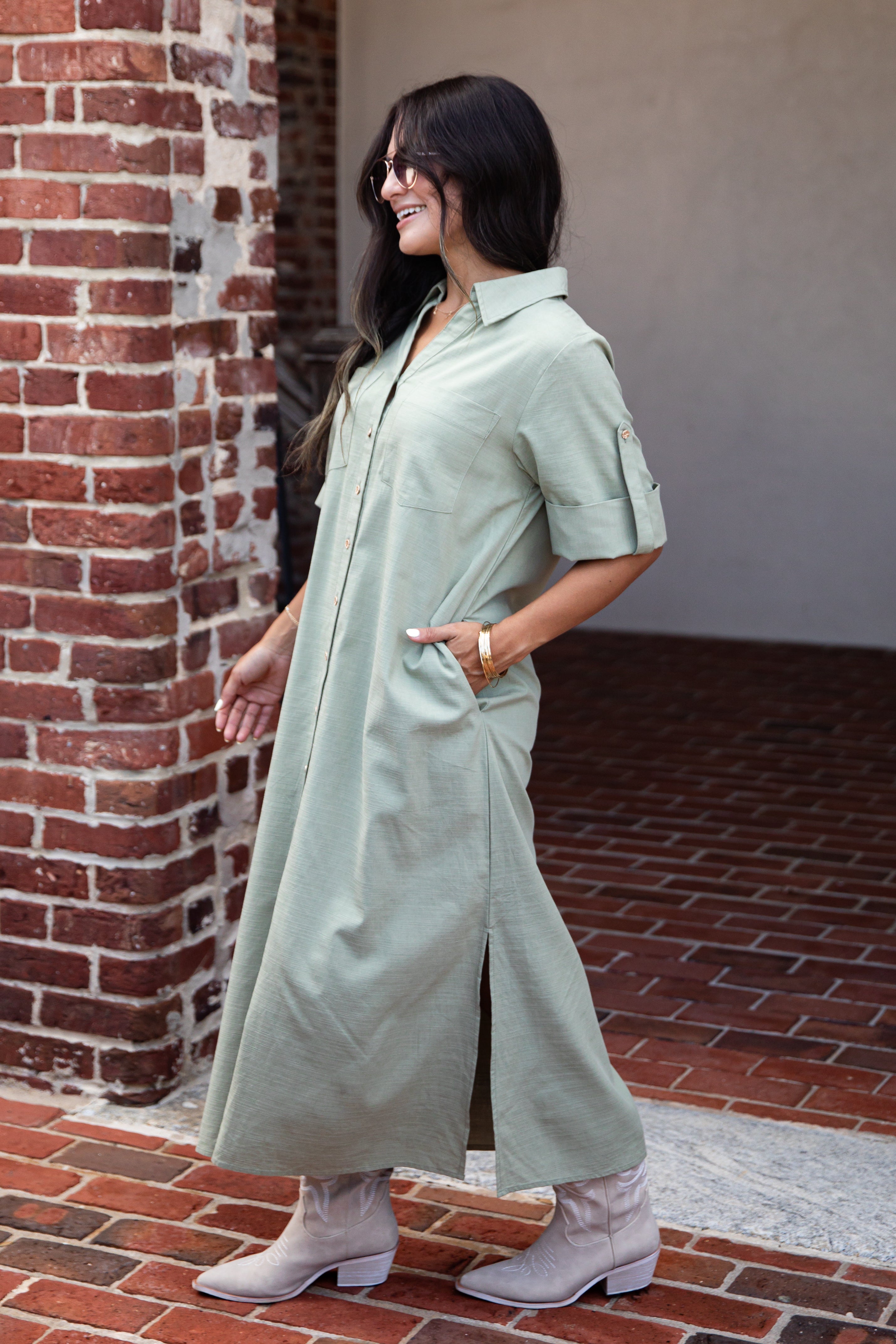 A woman wearing a sage green short-sleeve shirt dress with side slits and button-front closure, paired with light beige ankle boots, standing outdoors next to a red brick column.