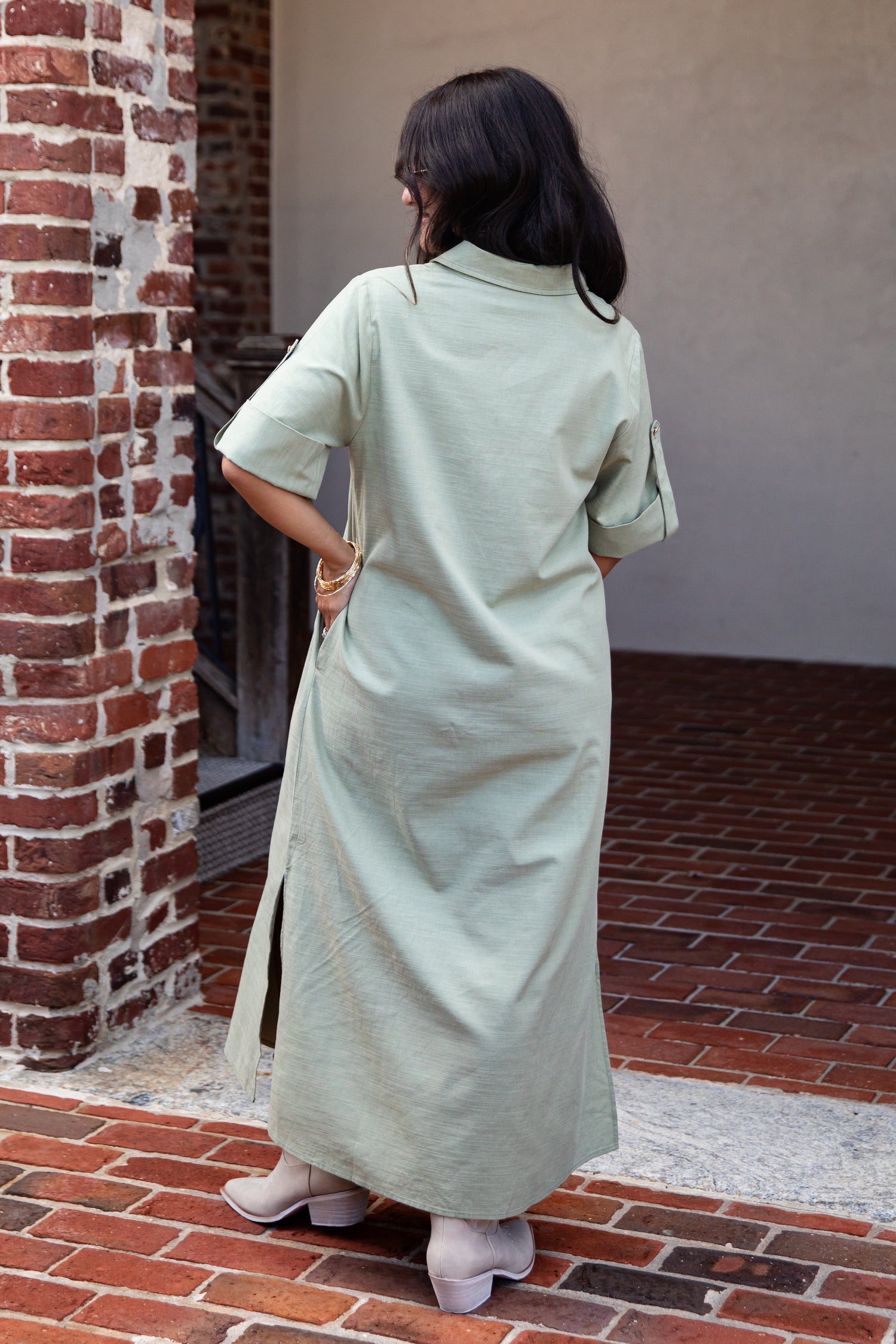 A woman viewed from behind wearing a light sage green linen shirt dress in an outdoor setting with a brick wall and red brick floor.