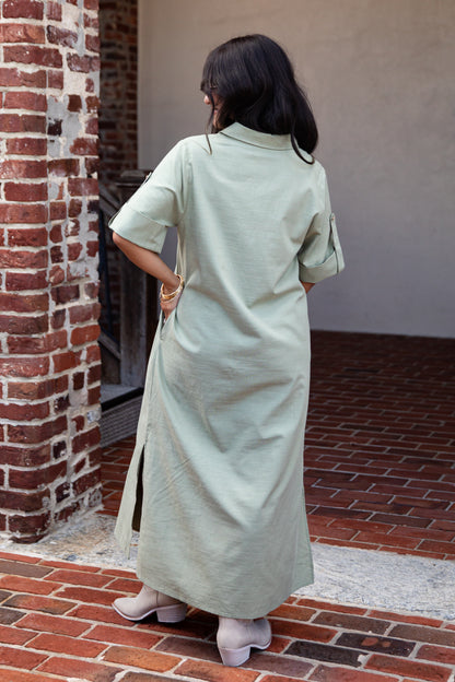 A woman viewed from behind wearing a light sage green linen shirt dress in an outdoor setting with a brick wall and red brick floor.