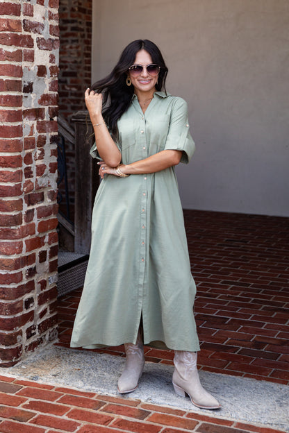 A woman wearing a sage green midi shirt dress and light beige boots stands outdoors in front of a brick column and wall.