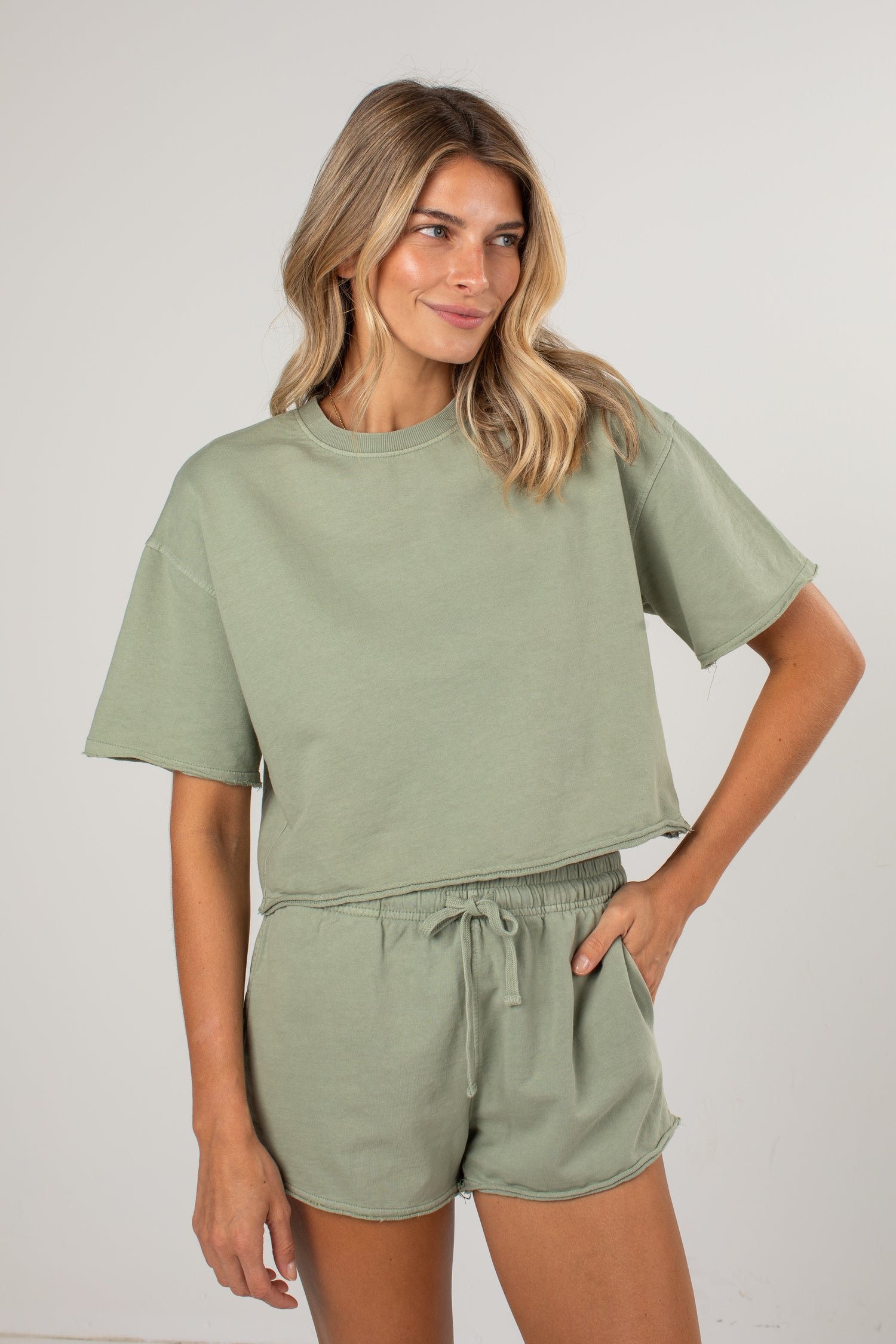 A woman modeling a matching sage green lounge set consisting of a short-sleeve cropped t-shirt and drawstring shorts, photographed against a neutral light gray studio background.