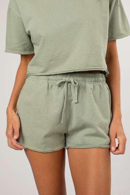 A person wearing a matching sage green lounge set consisting of a cropped short-sleeve top and drawstring shorts, shown from the chest down against a plain light gray background.