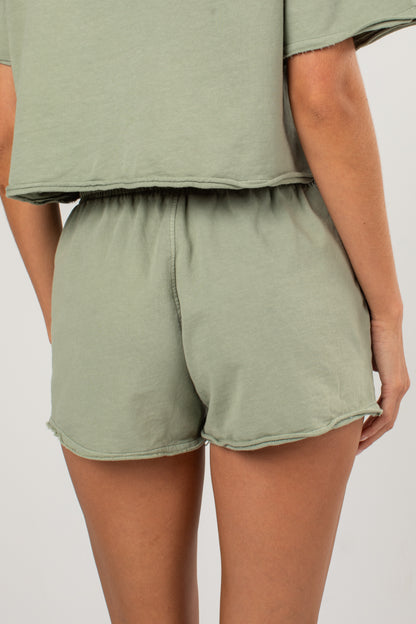 A person wearing a matching sage green lounge set, featuring a cropped short-sleeve top and high-waisted shorts with a raw hem, shown from the back against a light gray studio background.