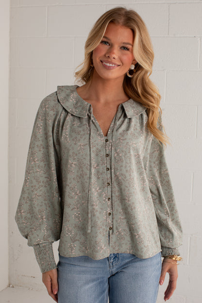A woman wearing a sage green long-sleeve blouse with a delicate pink floral pattern, a wide collar, and a button-down front, paired with blue jeans against a white brick wall.
