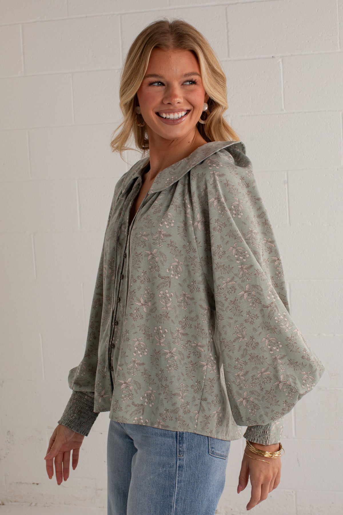 A smiling blonde woman wearing a sage green hooded poncho-style top with a subtle floral pattern and blue jeans, standing against a white painted brick wall.