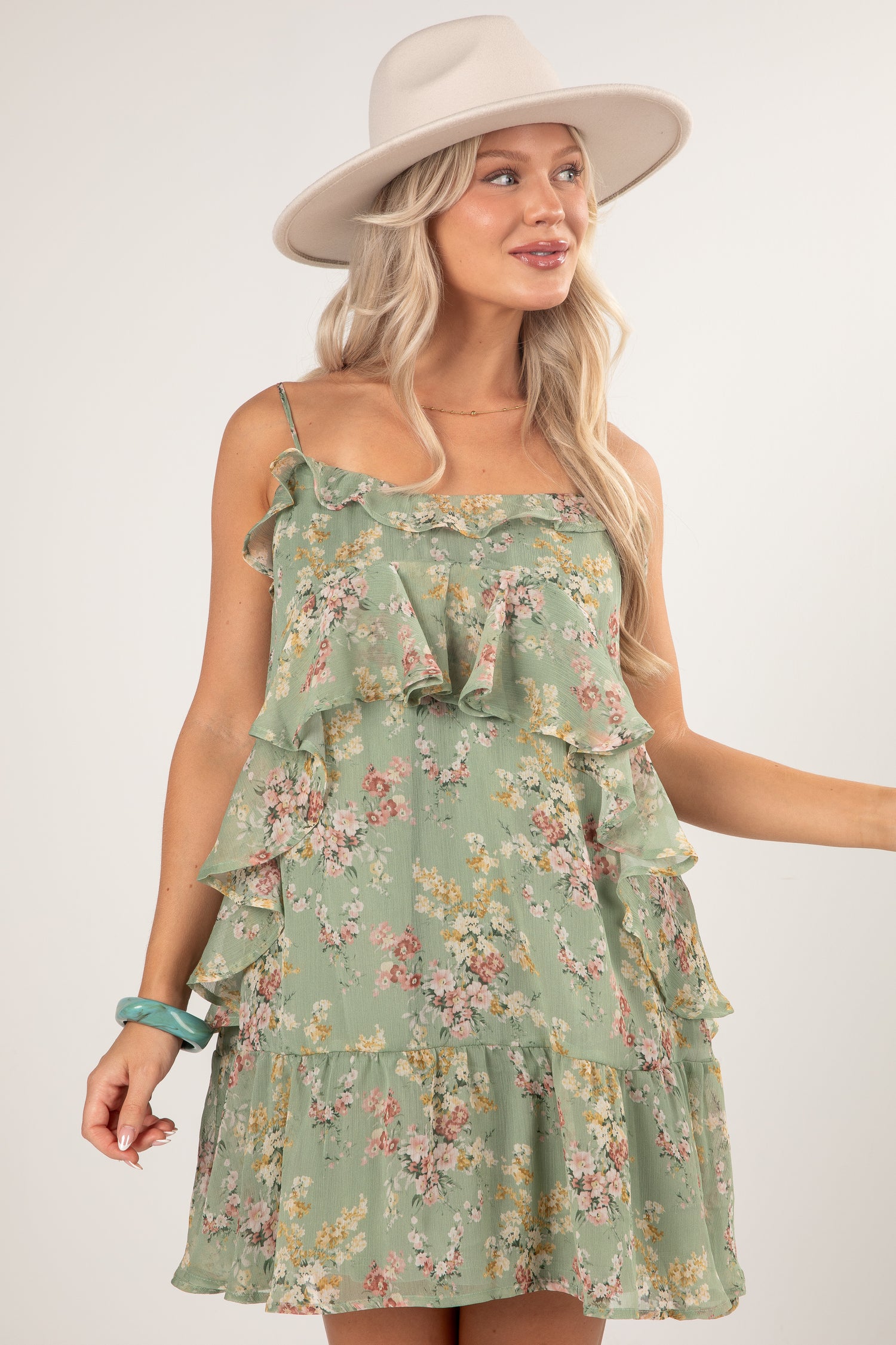 A woman wearing a sage green tiered ruffle mini dress with a pink and yellow floral print, styled with a beige wide-brim hat and a turquoise bracelet against a light gray studio background.