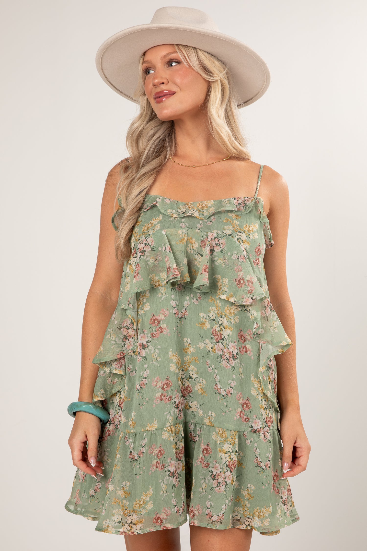 A woman wearing a sage green ruffled spaghetti-strap romper with a pink and yellow floral print, styled with a wide-brimmed cream hat against a light gray studio background.
