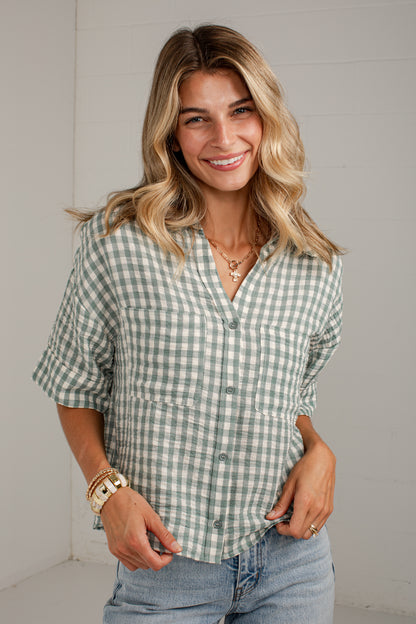 A smiling blonde woman wearing a sage green and white gingham short-sleeve button-up shirt with rolled cuffs, paired with light-wash blue jeans, standing against a white paneled wall.