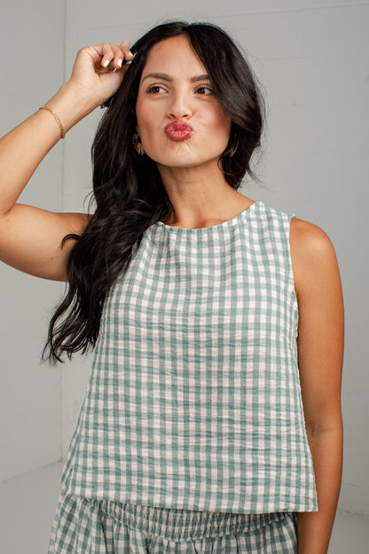 A woman wearing a sleeveless green and white gingham top, posing against a light gray studio background.