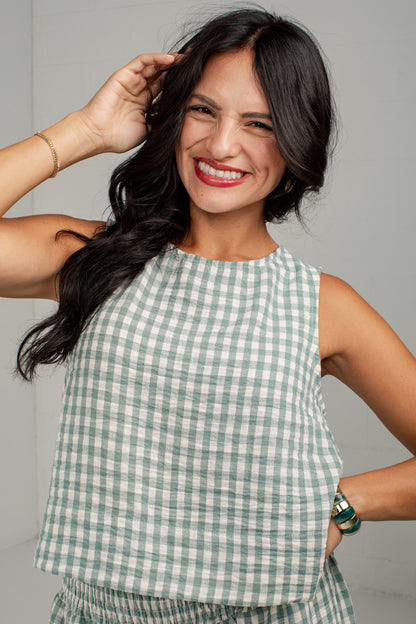 A smiling woman with long dark hair wearing a sleeveless light green and white gingham check top, posing against a neutral light gray background.