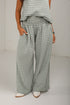 A person wearing wide-leg gingham pants in a muted sage green and white check pattern, paired with a matching top and white shoes, standing against a plain light gray wall.