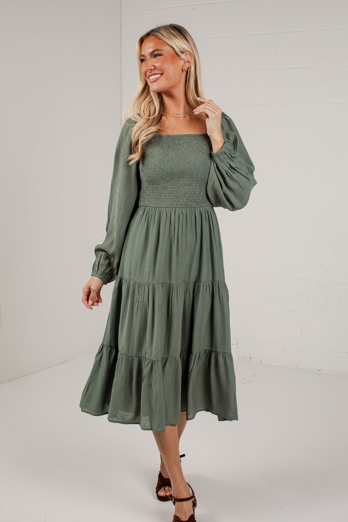 A woman modeling a sage green midi dress with long puff sleeves, a smocked bodice, and a tiered skirt, standing against a white brick wall background.