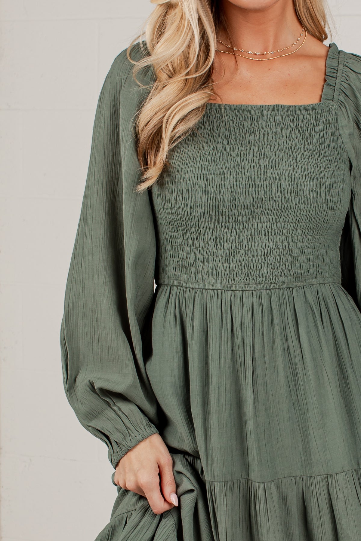 A woman wearing an olive green smocked dress with long balloon sleeves and a square neckline, shown from the shoulders to the hips against a light neutral background.