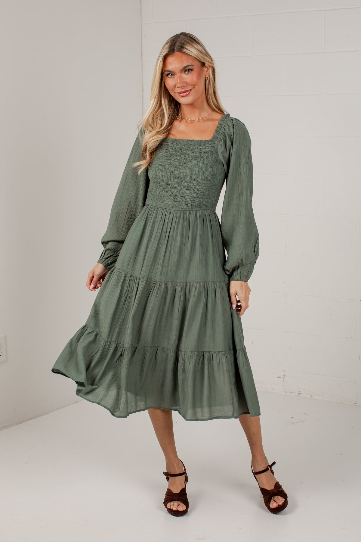 A woman modeling a sage green tiered midi dress with long puff sleeves and a square neckline, paired with brown strappy heels, in a bright white studio setting.