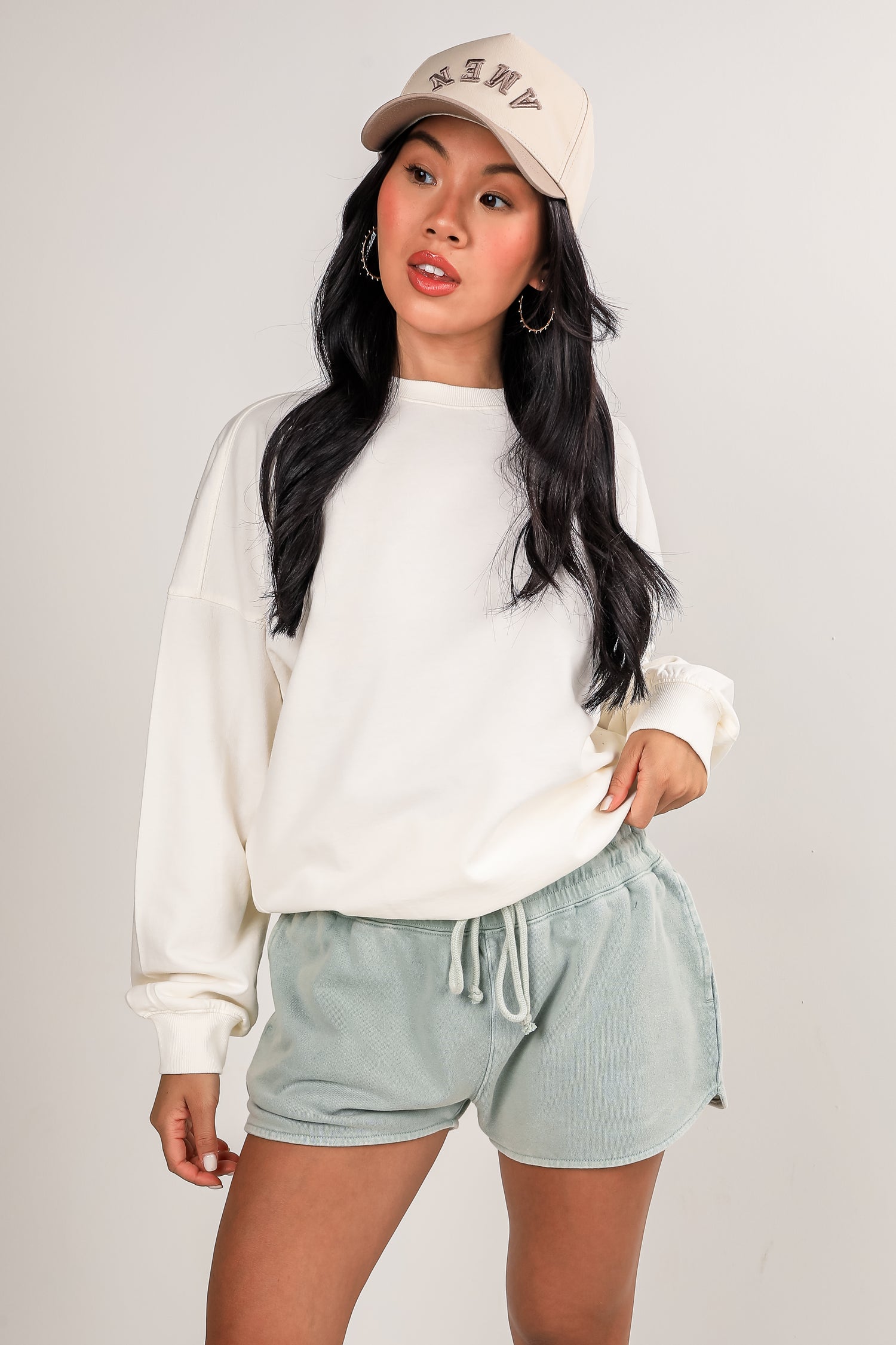 Woman wearing a white sweatshirt and light green shorts with a beige cap on a plain background