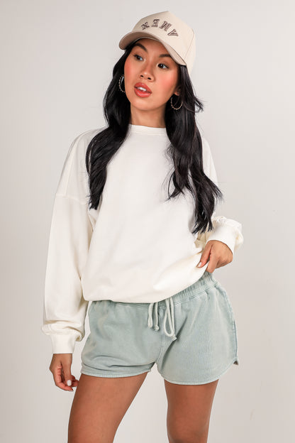 Woman wearing a white sweatshirt and light green shorts with a beige cap on a plain background