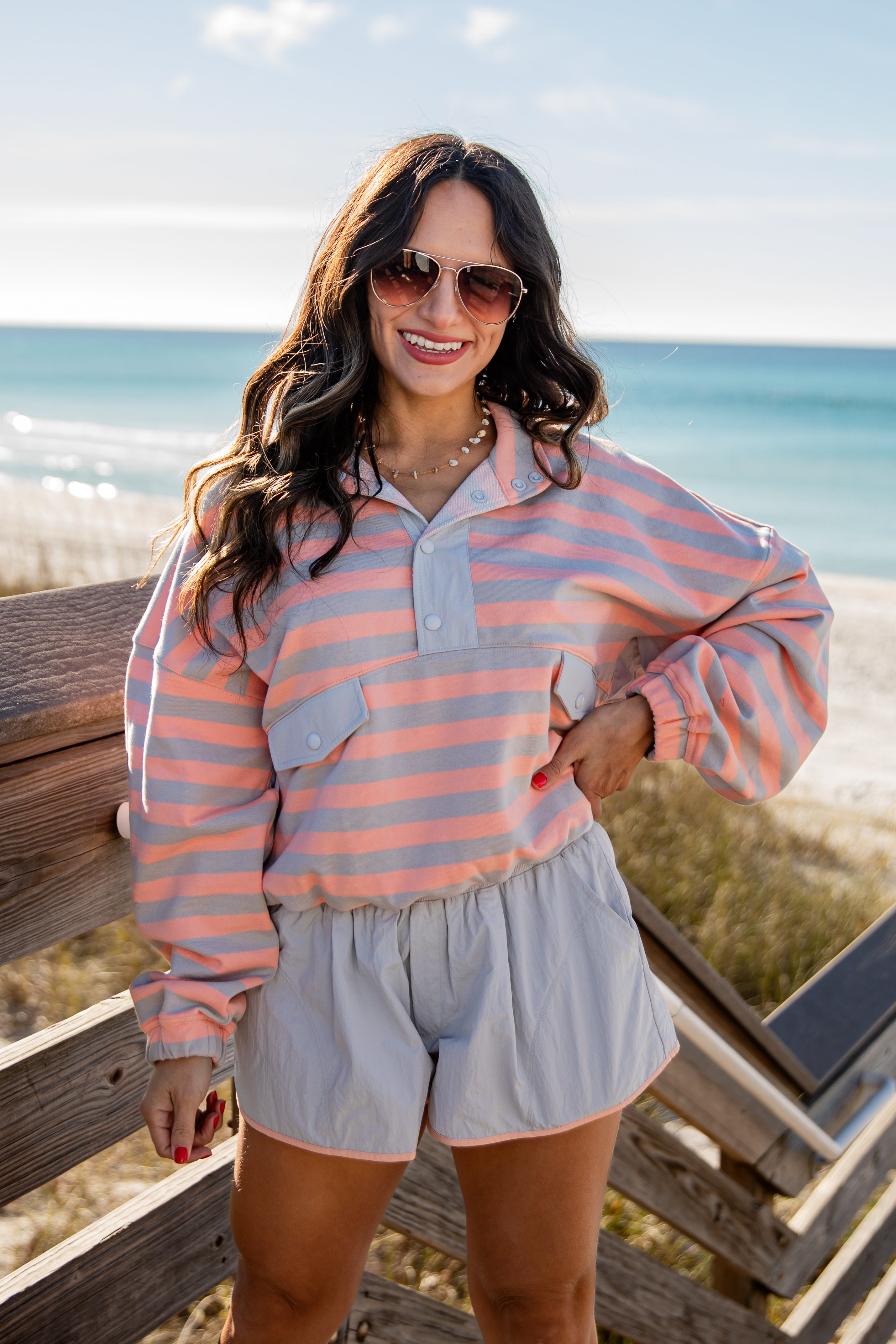 Woman wearing a striped shirt and shorts standing on a wooden deck by the beach.