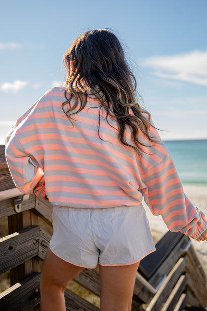 Person wearing a striped shirt and shorts standing on a wooden deck by the beach.