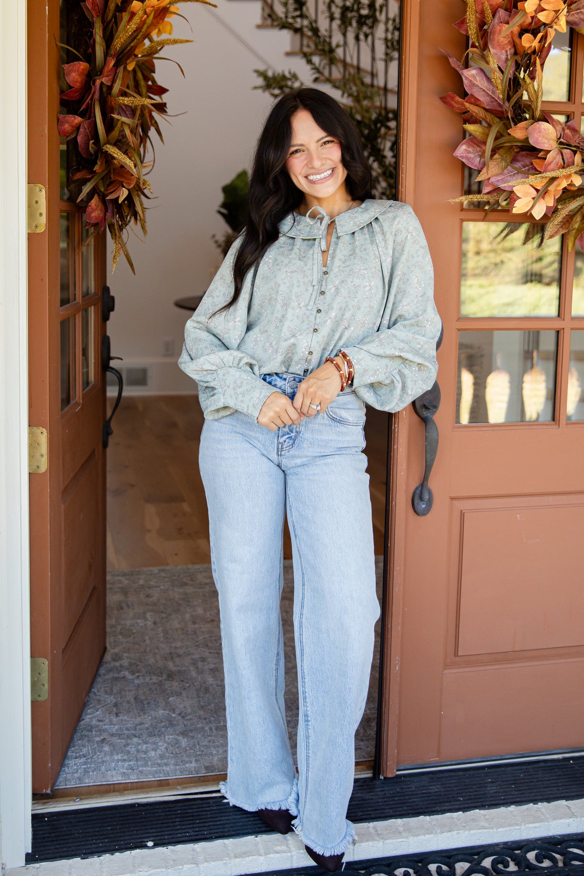 A woman stands in a doorway wearing a light sage green hooded button-up shirt and light blue wide-leg jeans. The setting features a rustic brown door with glass panes, fall wreaths, and a gray rug.
