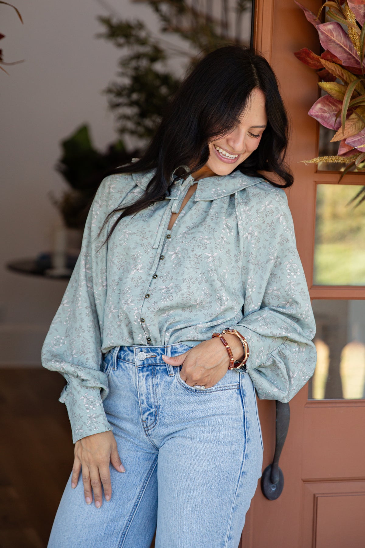 A woman wearing a light sage green long-sleeve blouse with a subtle floral pattern and light-wash blue jeans, standing in a warmly lit indoor setting.