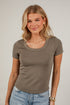 A woman with blonde hair wearing a fitted short-sleeve scoop-neck t-shirt in a muted taupe-brown color, paired with light-wash blue jeans, standing against a white brick wall.