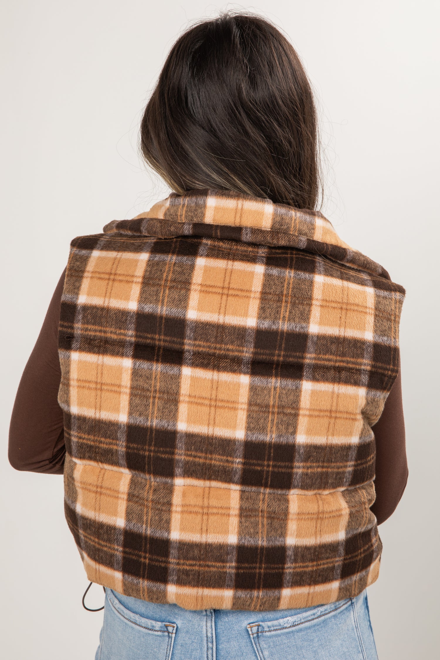 Person wearing a plaid vest over a brown shirt and blue jeans on a white background