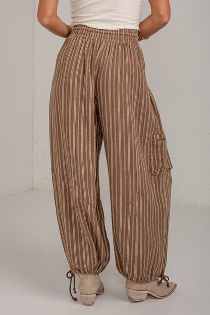 A person shown from the back wearing loose brown and beige vertically striped cargo pants with drawstring hems and light beige heeled sandals, standing against a plain light gray wall.