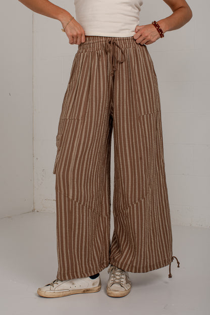 A person wearing brown and beige vertically striped wide-leg pants with a drawstring waist, paired with a white top and white sneakers, standing against a plain light gray wall.
