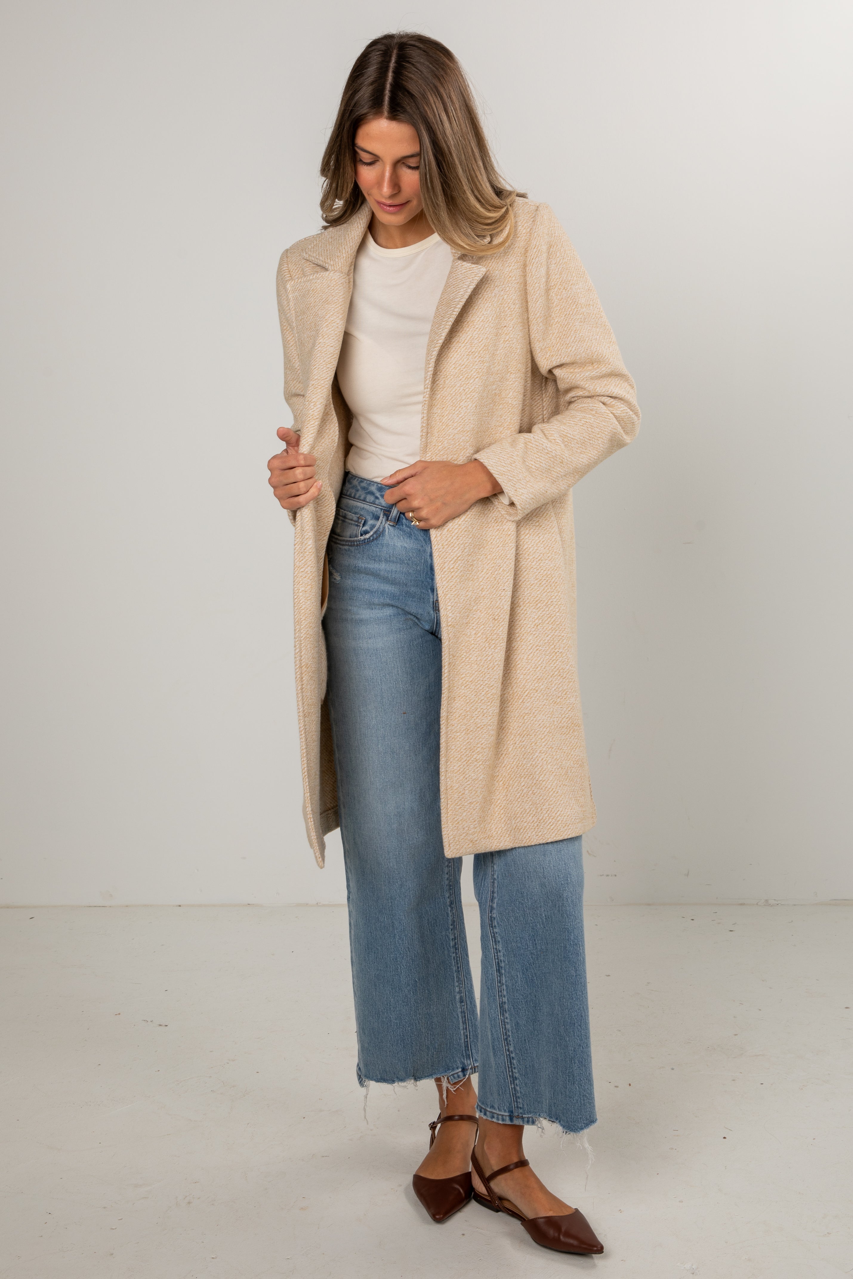 Woman wearing a beige coat, white shirt, and blue jeans on a plain background
